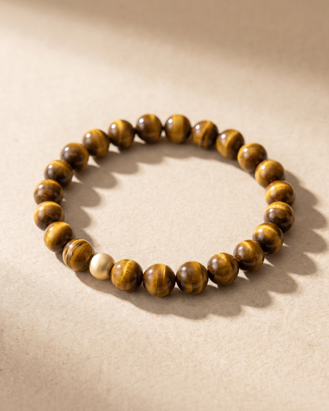Pulsera de ojo de tigre con cuenta dorada, tumbada en plano sobre fondo arena