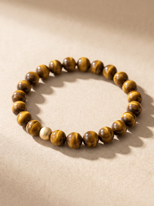 Pulsera de ojo de tigre con cuenta dorada, tumbada en plano sobre fondo arena